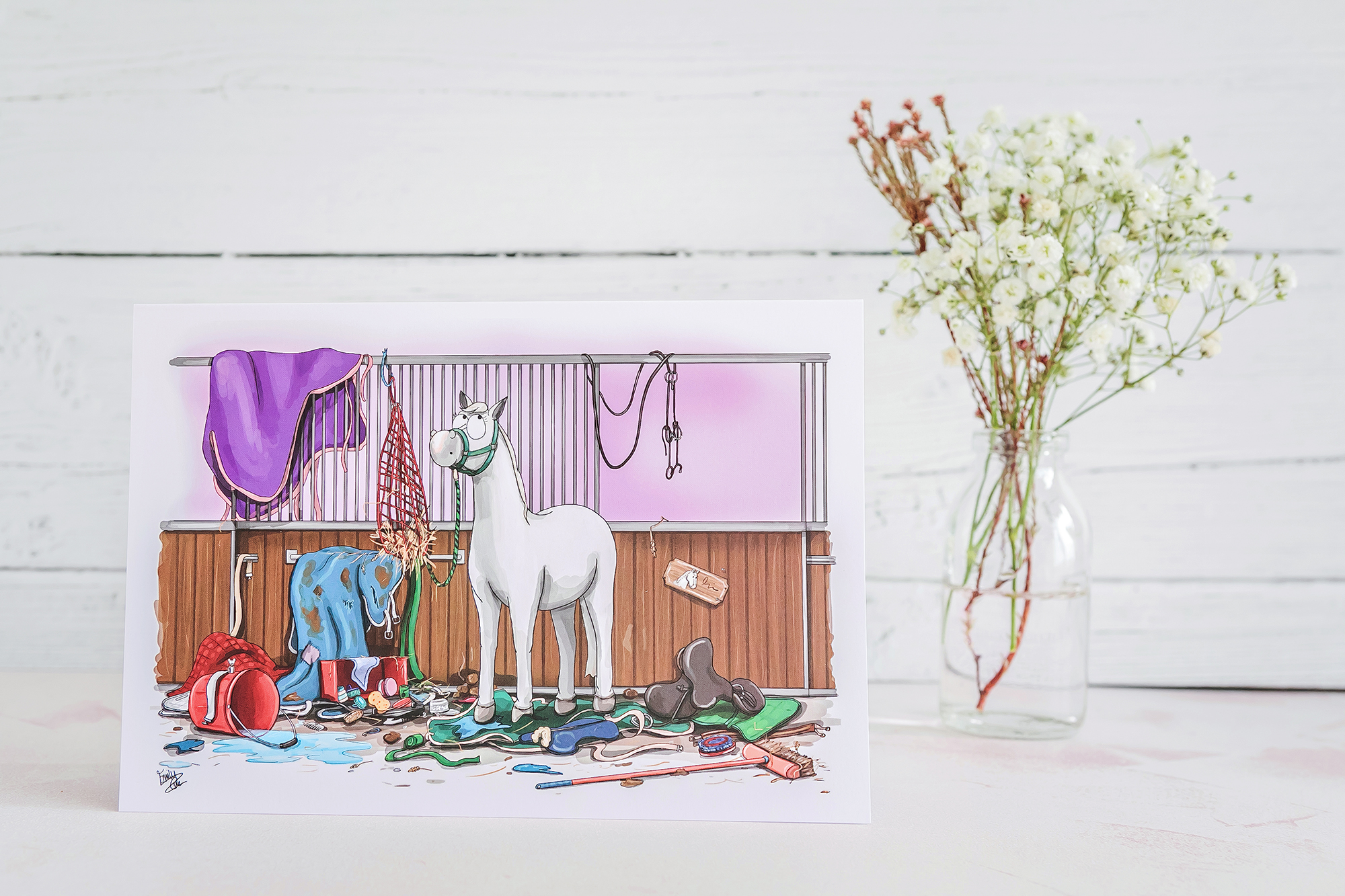 A5 Greeting card next to flowers. Cad illustrates a design of a white horse standing in a pile of rugs and broken items.