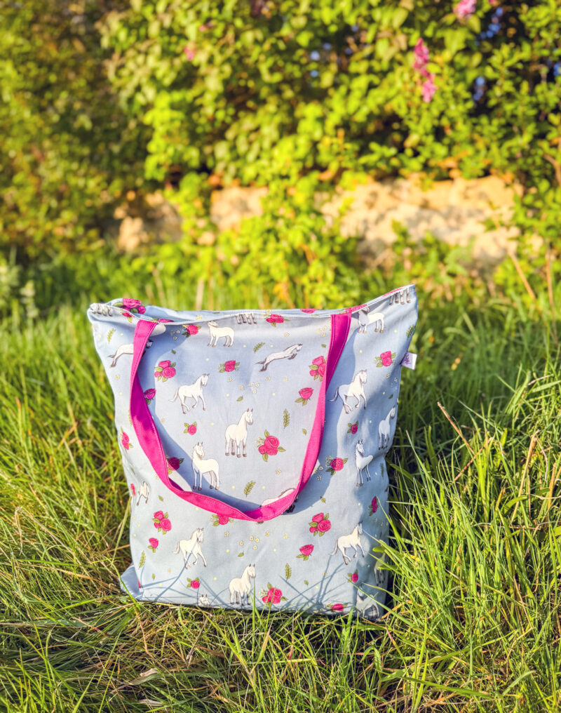 Grey horse patterned tote bag with pink rose design and magenta handles sitting in long grass