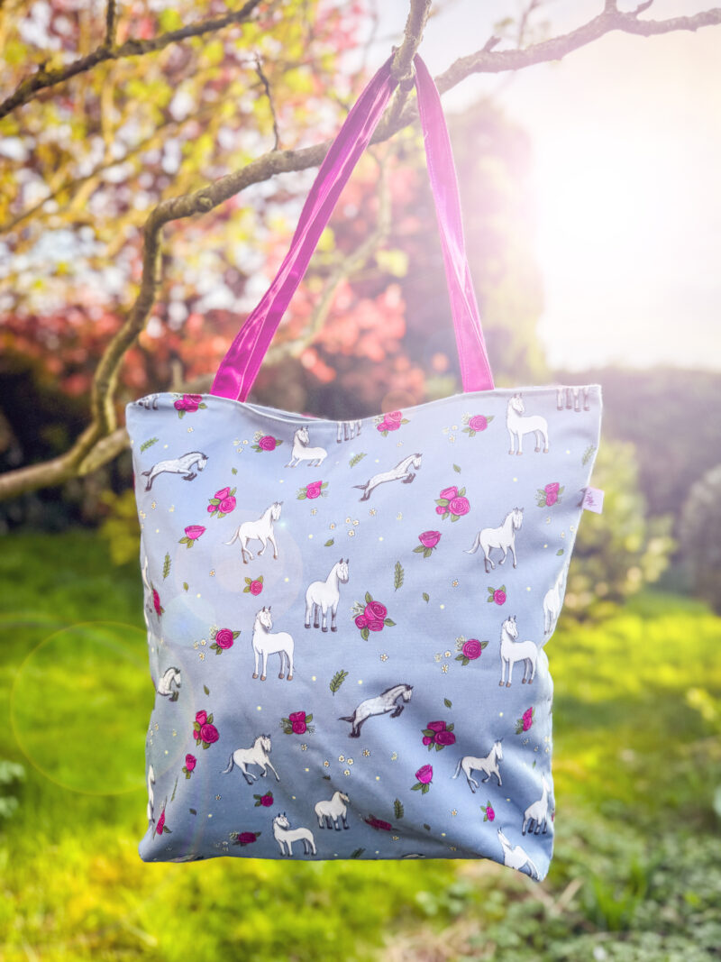Grey horse and rose patterned tote bag hanging from tree with sunlight and pink handles